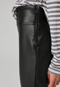 Pantaloni in pelle nera con zip laterale, caratterizzati da una texture liscia e un design aderente, abbinati a una maglietta a righe grigio chiaro.