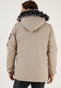 Beidzis beigu apvalks ar pelēku mākslīgo ādu apdari uz kapuces, ar teksturētu audumu, sānu kabatām un pieguļošu siluetu, skatīts no aizmugures.