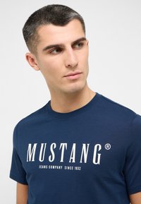 Jūras zilas kokvilnas T-krekls ar apaļu kakla izgriezumu un īsām piedurknēm. Priekšpusē ir izteiksmīgs balts teksts "MUSTANG" un "JEANS COMPANY SINCE 1932".
