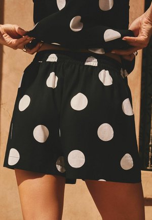 Hände heben ein schwarzes Oberteil an und zeigen passende schwarze Shorts mit großen weißen Punkten, getragen im Freien bei warmem Sonnenlicht.