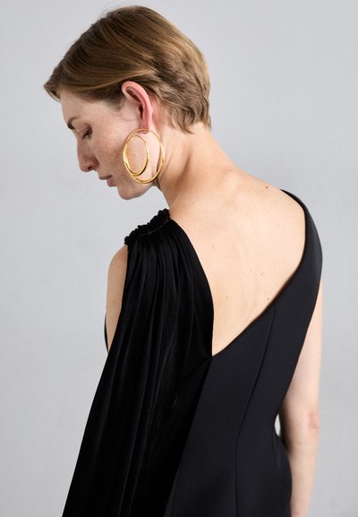 Juoda suknelė su asimetriniu dizainu, kurioje yra šilkinis drapiruotas rankovė ir prigludusi liemenė. Auksinės žiedinės auskarai užbaigia įvaizdį.