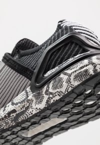 Zapato deportivo en negro y gris que presenta una parte superior de tejido texturizado, un acento de plástico negro y una suela con estampado de serpiente en blanco y negro.