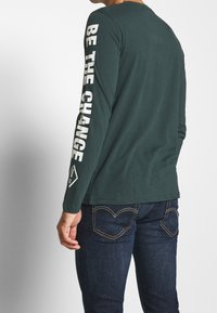 Man wearing dark green long-sleeve shirt with white "BE THE CHANGE" text on right arm, paired with dark blue jeans with visible stitching.