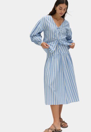 Vrouw die een blauw-wit gestreepte jurk met lange mouwen, V-hals en gestrikte details aanpast, met bruine platte sandalen tegen een witte achtergrond.