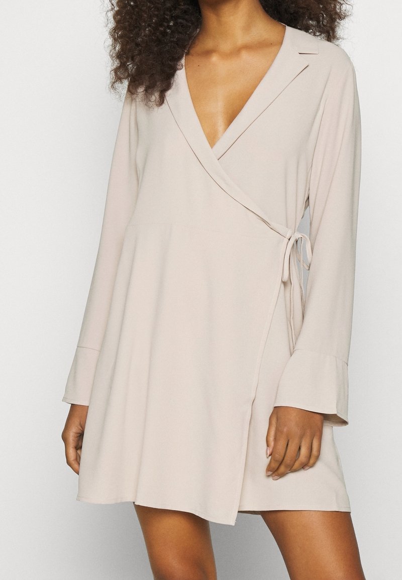 Femme portant une robe longue à manches longues beige avec un lien sur le côté et des poignets évasés, montrant une longueur mi-cuisse et un col en V.