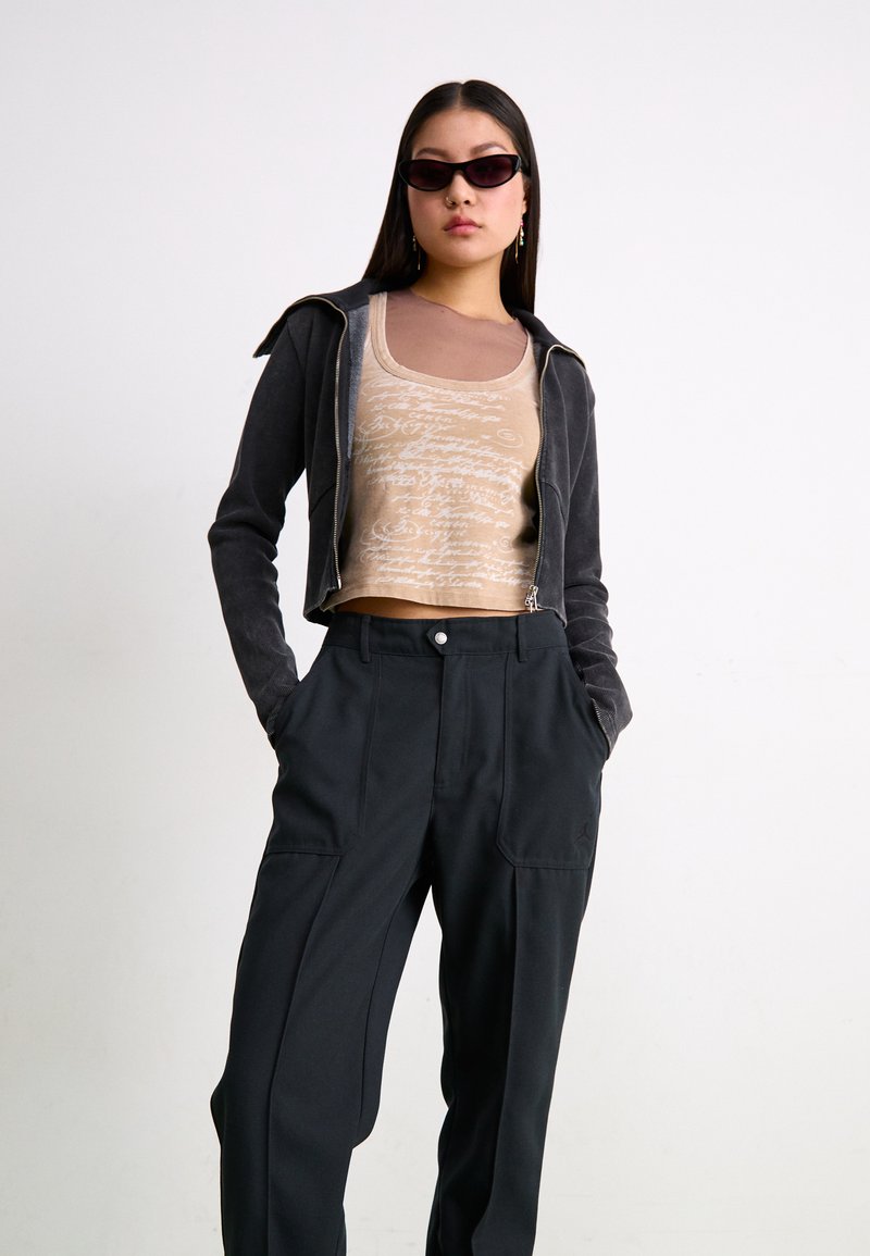 Graue Zip-Jacke über einem beigen Tanktop mit Schriftmuster; kombiniert mit schwarzen, locker sitzenden Hosen mit Seitentaschen. Schwarze Sonnenbrille.