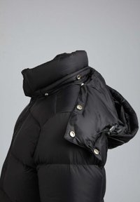 Schwarze Pufferjacke mit hohem Kragen, Druckknopfverschlüssen und verstellbarem Zugband. Strukturierter Stoff in einem gesteppten Muster.