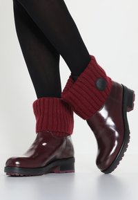 Botas de tobillo de cuero burdeos con puño de punto burdeos. El puño cuenta con un botón negro. Suela negra con patrón de tracción texturizado.