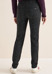 Persoon gekleed in donkergrijze textuurbroek en witte sneakers met bordeauxrode accenten, staand tegen een effen lichte achtergrond.