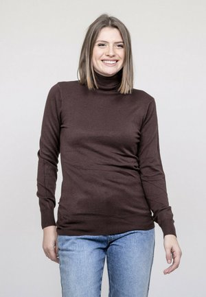 Femme souriante aux cheveux raides mi-longs porte un pull à col roulé marron et un jean bleu clair sur un fond uni.