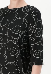 Svart topp med et hvitt blomstermønster og konturerte detaljer. Korte ermer og klassisk rund hals. Litt avslappet passform.