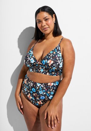 Kvinde med mørkt, mellemlangt hår iført en sort, blomsterprintet to-delt badedragt, stående mod en hvid væg med skygge.