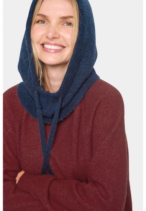 Femme souriante portant un bonnet en laine bleu et un pull bordeaux, les bras croisés devant un fond blanc uni.