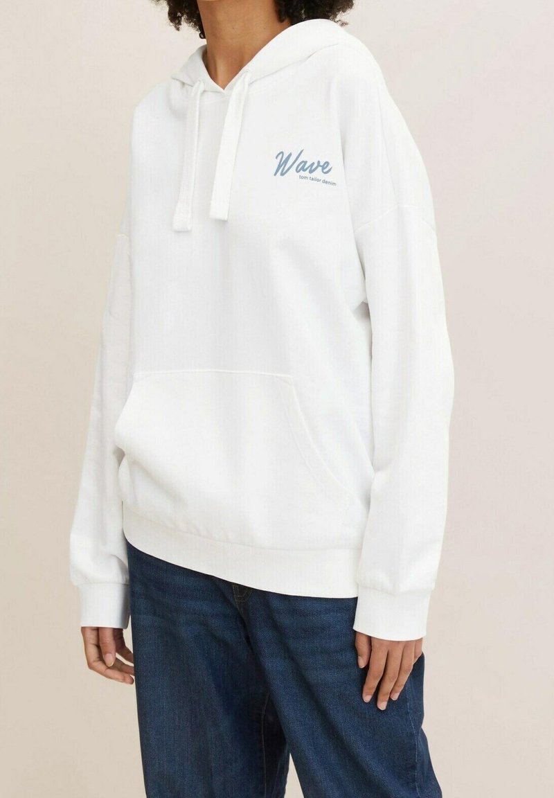 Persona vestida con una sudadera blanca que lleva el texto "Wave tom tailor denim" en el pecho y jeans azules, de pie contra un fondo beige liso.