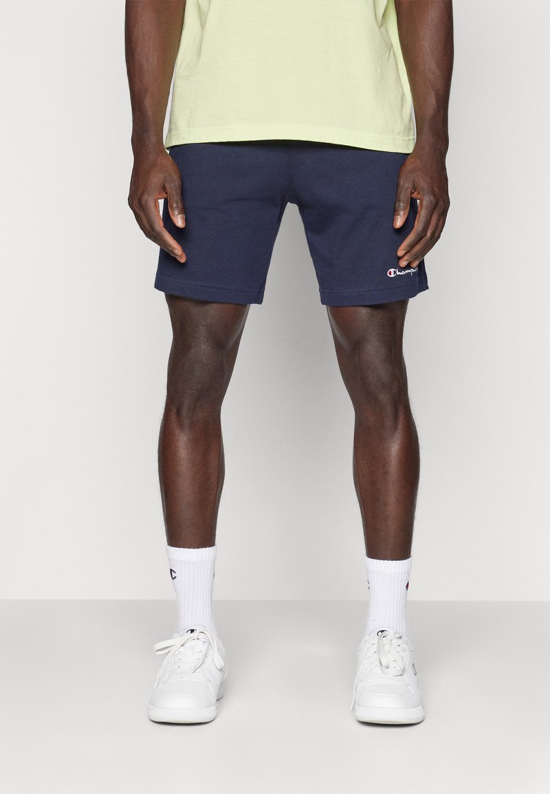 Pantalones cortos deportivos azul marino con cinturilla elástica, que presentan un pequeño parche de logo rojo y blanco. Combinados con zapatillas blancas y calcetines.