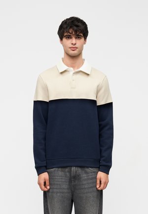 Jonge man met krullend donker haar, gekleed in een crème en marineblauw poloshirt met lange mouwen en verweerde grijze spijkerbroek, staand tegen een witte achtergrond.