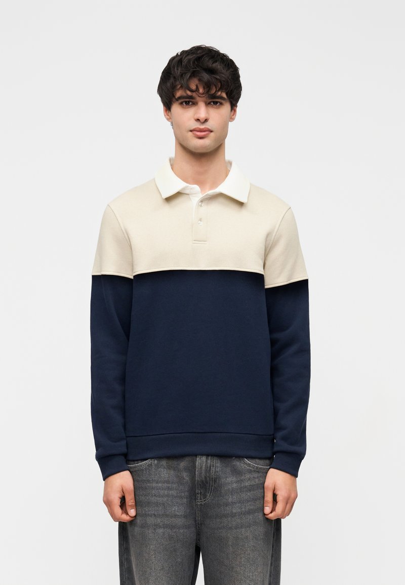 Νέος άνδρας με σγουρά σκούρα μαλλιά, φορώντας πόλο με μακριά μανίκια σε κρεμ και ναυτικό μπλε και ξεθωριασμένα γκρι τζιν, στέκεται μπροστά σε λευκό φόντο.