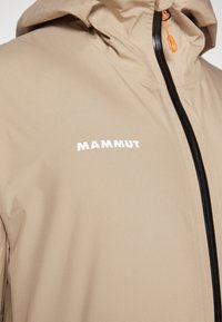 Beige lättviktsjacka med huva och strukturerad yta, med framzip och vit "MAMMUT" logotyp på bröstet.
