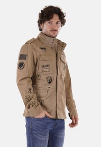 Giacca militare color sabbia con diverse patch, tra cui una bandiera americana e vari distintivi, dotata di colletto e bottoni frontali.