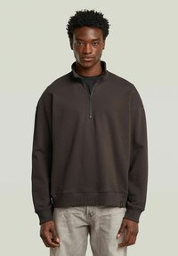 Bruine sweatshirt met een hoge kraag, halve ritssluiting aan de voorkant en geribbelde manchetten. Heeft een relaxte pasvorm en een zachte textuur. Draagt bij lichte grijze broek.