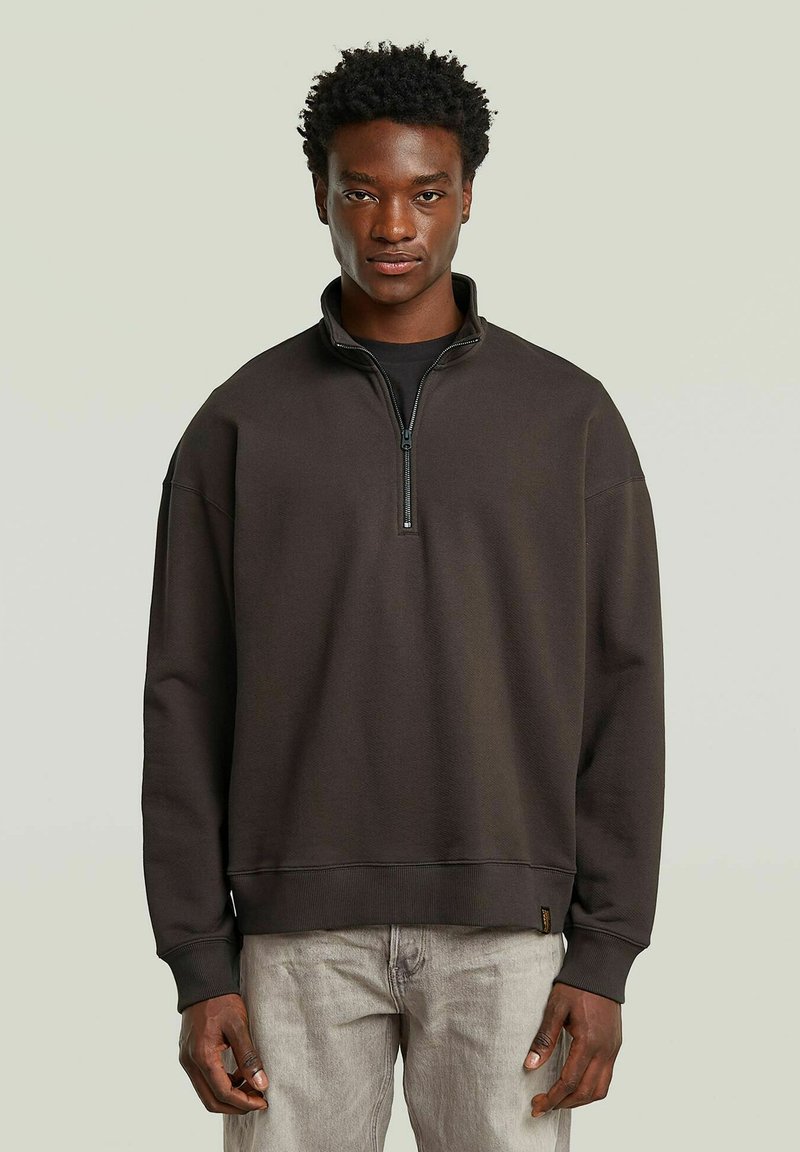 Bruine sweatshirt met een hoge kraag, halve ritssluiting aan de voorkant en geribbelde manchetten. Heeft een relaxte pasvorm en een zachte textuur. Draagt bij lichte grijze broek.