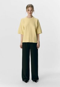 Haut jaune à manches courtes avec un col rond, associé à un pantalon noir à jambe large et des chaussures noires à bouts pointus, porté sur un fond neutre.