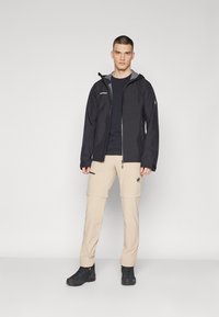 Veste étanche noire avec doublure grise, pantalon beige à zip détachable, et chaussures de randonnée noires. Comprend un logo sur la veste et des poches sur les cuisses du pantalon.