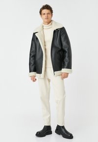 Giacca di pelle nera con colletto e polsini in shearling bianco, dotata di chiusura a zip e vestibilità morbida, abbinata a pantaloni color crema e stivali neri.