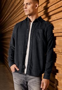 Camisa negra de botones en una tela texturizada, con un bolsillo en el pecho y un corte recto, llevada sobre una camiseta blanca con jeans grises.