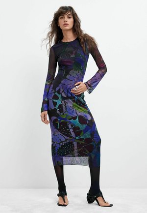 Frau trägt ein langärmeliges, knielanges Kleid mit abstrakten blauen, grünen und violetten Mustern, schwarze Strumpfhosen und schwarze Sandalen mit Absatz.