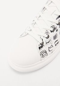 Bílé tenisky s texturovaným svrškem, na kterém jsou černé grafické designy obličeje, plochými bílými tkaničkami a zaoblenou gumovou podrážkou.