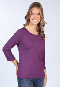 Lila 3/4-Ärmeloberteil aus weichem Stoff mit rundem Ausschnitt, tailliertem Design und seitlichen Schlitzen. Kombiniert mit blauen Jeans.