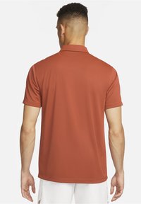 Homme aux cheveux courts portant un polo à manches courtes couleur rouille et un short blanc, vu de dos sur un fond blanc.