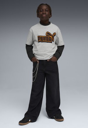 Dziecko stoi z rękami w kieszeniach, ubrane w szarą koszulkę Puma na czarnej bluzie z długim rękawem, czarne szerokie spodnie z łańcuchami i czarne trampki.