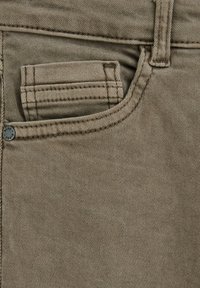 Beige denimstof med en frontlomme med syningsdetaljer, en metalknap med indgraveret "BLUE" og en glat teksturoverflade.