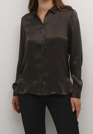 Kobieta w ciemnobrązowej satynowej koszuli na długi rękaw zapinanej na guziki, w zestawieniu z czarnymi spodniami, stojąca na tle jednolitego jasnego tła.