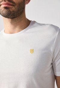Fehér pamut póló kör alakú nyakkivágással, a mellrészen egy kis sárga, hímzett emblémával. Sima textúra, rövid ujjú.