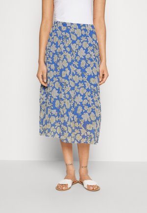Femme portant une jupe midi bleue avec des motifs floraux beiges, un haut blanc, des sandales blanches, debout devant un fond blanc uni.