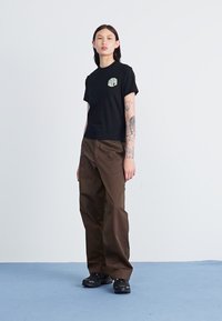 Svart kortermet t-skjorte med et lite grafisk design, kombinert med løstsittende brune bukser og svarte joggesko.