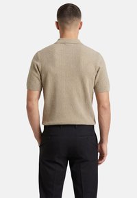 Homme debout, de dos, portant un polo en maille beige à manches courtes et un pantalon noir, sur un fond blanc uni.