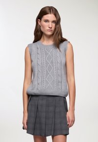 Maglione grigio lavorato a maglia senza maniche con motivo a trecce, collo e orlo a coste, abbinato a una gonna grigia a quadri. Dettaglio della trama del tessuto visibile.