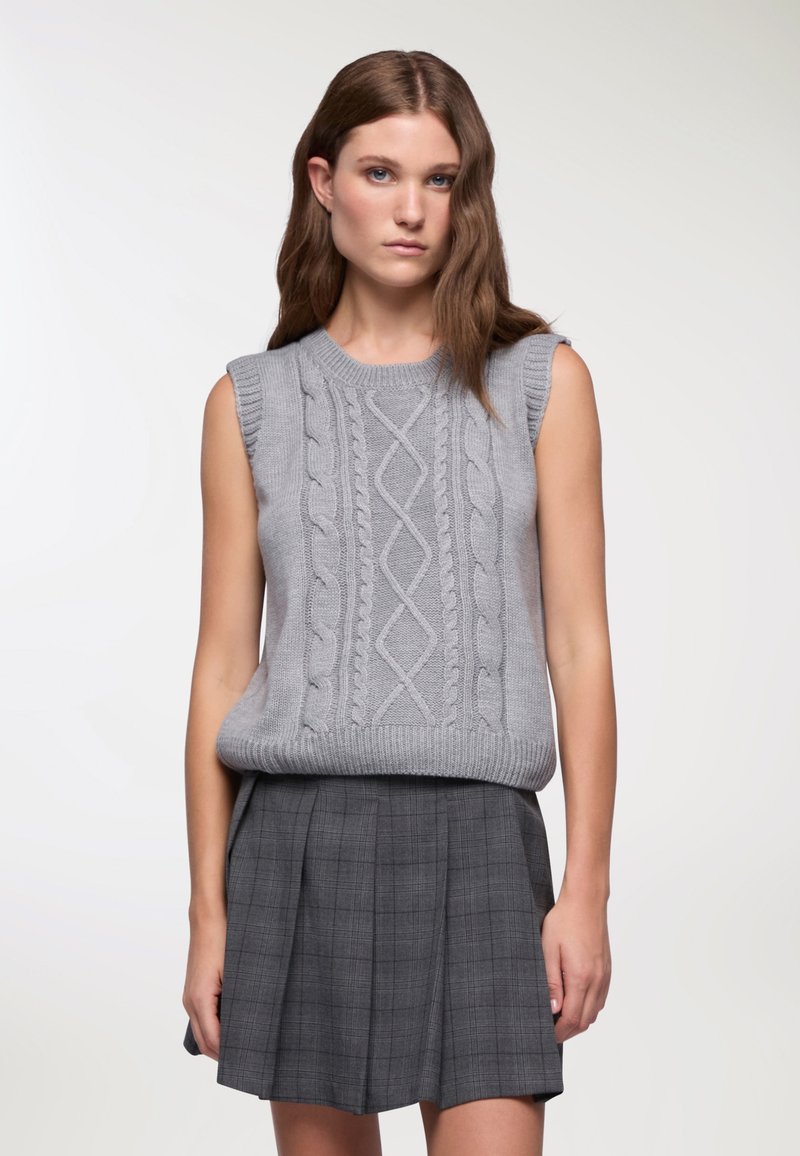 Maglione grigio lavorato a maglia senza maniche con motivo a trecce, collo e orlo a coste, abbinato a una gonna grigia a quadri. Dettaglio della trama del tessuto visibile.