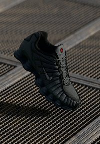 Shoe desportivo Nike preta com sola segmentada única e parte superior com padrão em grelha, exposta acima de uma superfície de grelha de metal texturizada.