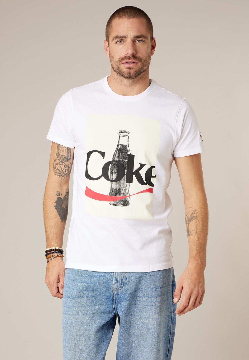 Homme aux cheveux courts et tatouages, portant un t-shirt blanc avec un graphisme vintage de bouteille de Coke et un jean bleu clair, debout devant un fond uni.