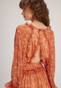 Langärmliges Kleid in Orange, mit einem Rüschenausschnitt, offenem Rücken und einem wellenförmigen Muster. Leichter, strukturiert Stoff.