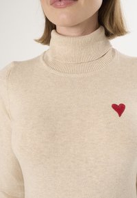 Maglione beige a collo alto realizzato in tessuto morbido, con un piccolo ricamo a forma di cuore rosso sul lato sinistro del petto. Collo e polsini a costine.