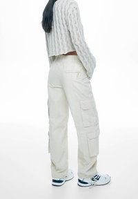 Pantalon cargo crème avec multiples poches, taille élastique et coupe décontractée, associé à un pull court en maille texturée blanche et des baskets.