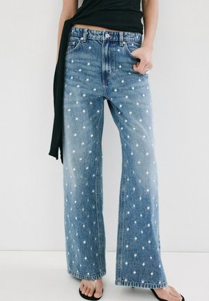 Persona con jeans azules de tiro alto y pierna ancha con lunares blancos, combinados con una camiseta negra y sandalias negras tipo tanga, de pie contra una pared blanca.