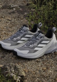 Zapatos deportivos grises con una parte superior texturizada, que cuentan con rayas blancas, punteras reforzadas y una suela acolchada para mayor soporte.