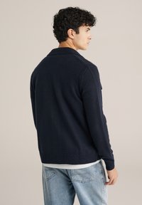 Marinblå stickad kofta med krage och ribbade ärmar. Har en strukturerad yta och subtila sömdetaljer.